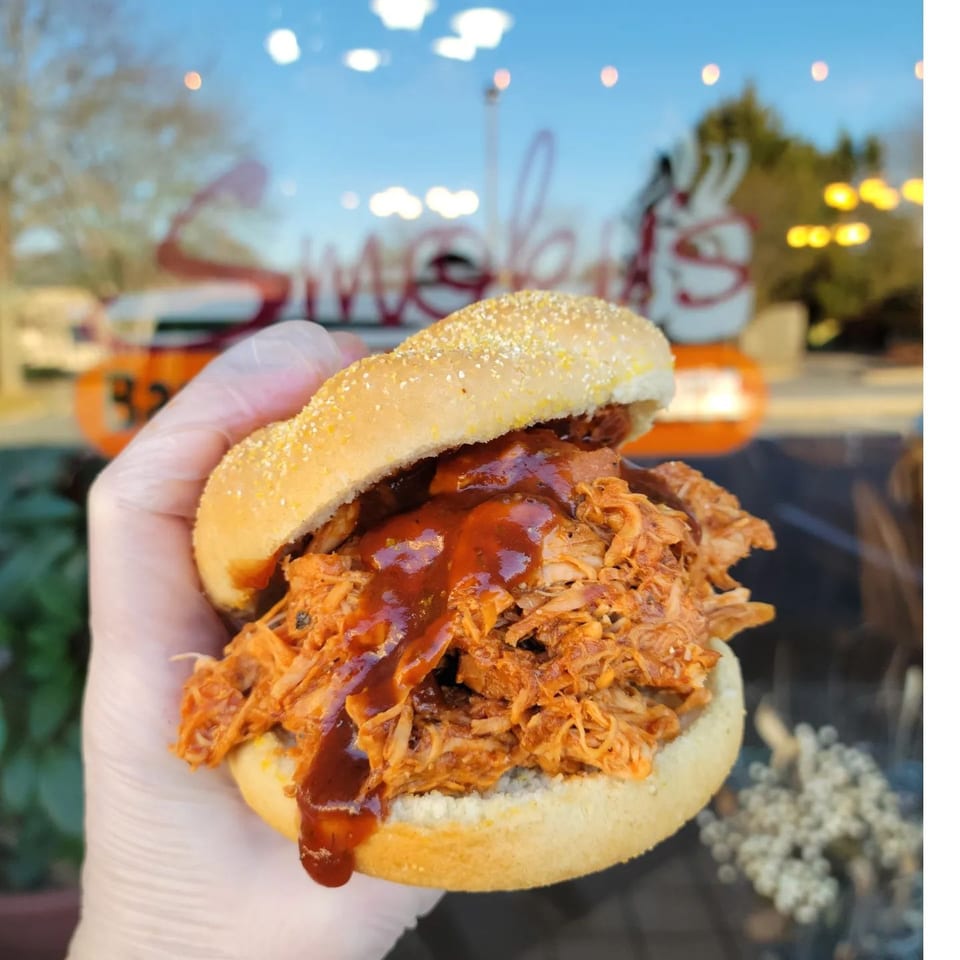 Gloved hand holding a pulled pork sandwich with barbecue sauce, storefront visible in the background.