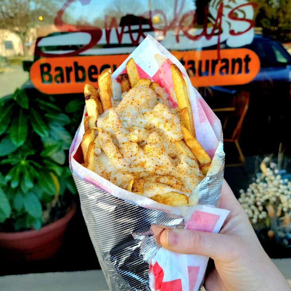 Hand holding a cone of cheesy fries with seasoning, in front of a restaurant window.