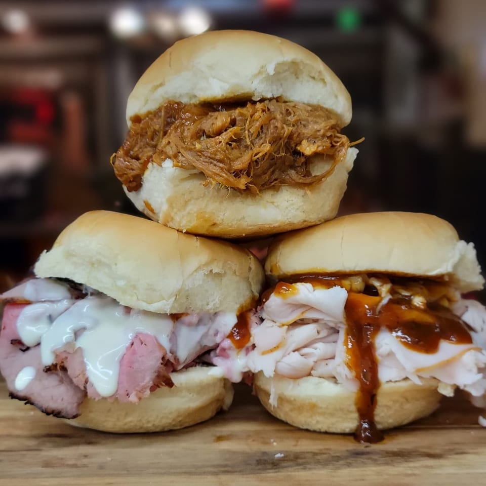 Three sliders on a wooden board: pulled pork, roast beef with white sauce, and turkey with BBQ sauce.