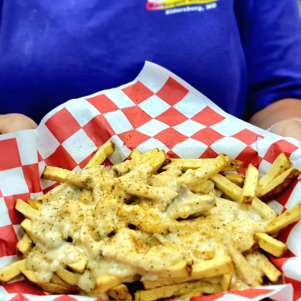 A person in a blue shirt holds a basket of Smokey's Barbecue fries topped with creamy cheese sauce and seasoning.