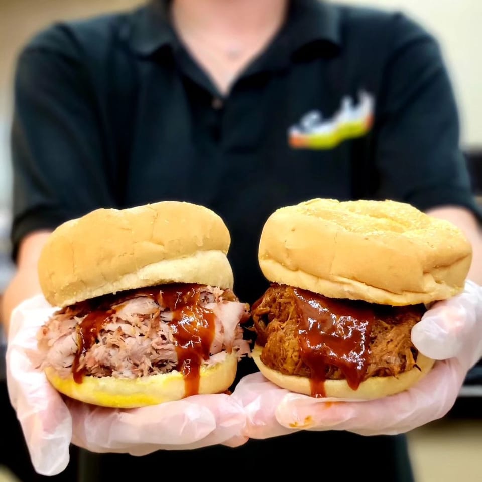 Gloved hands hold two barbecue sandwiches, one with sliced meat and one with pulled pork.