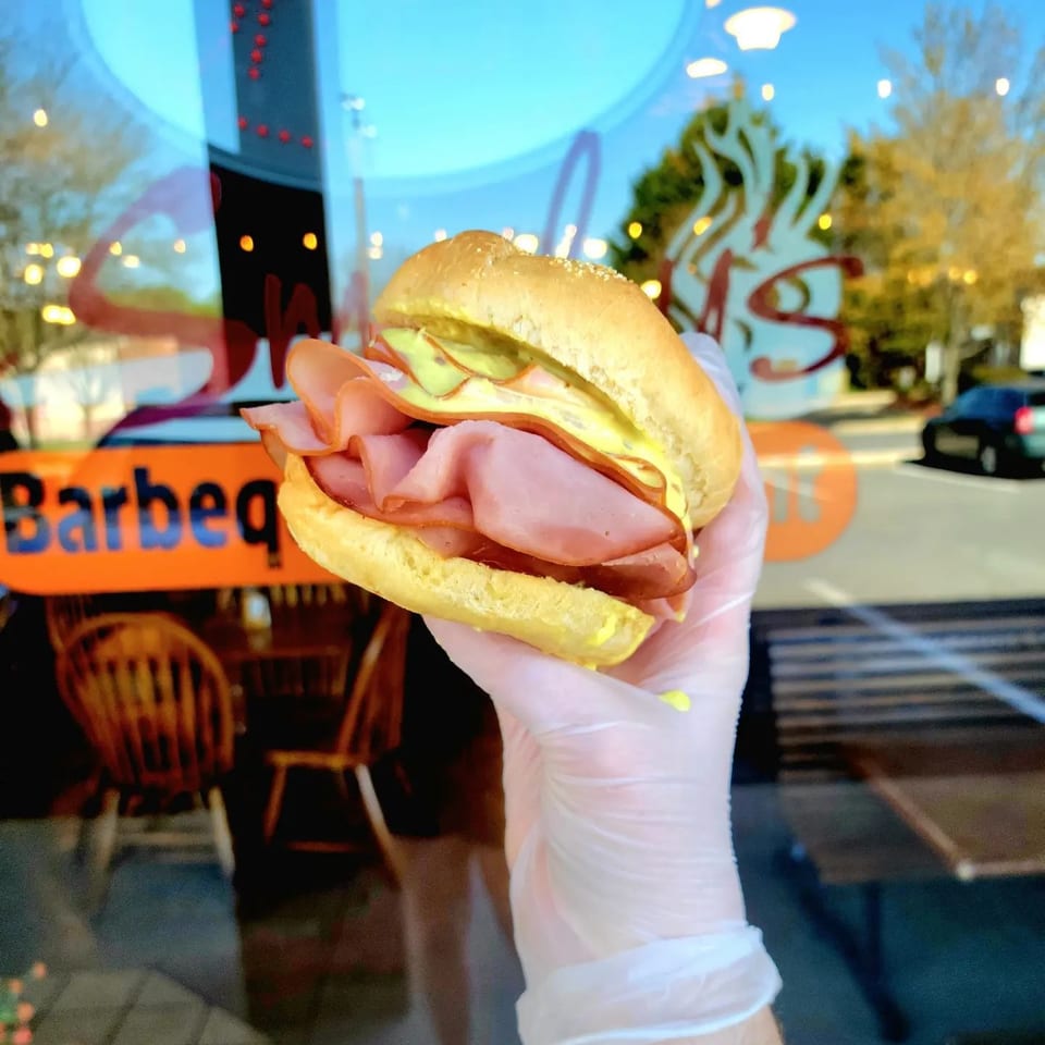 Gloved hand holding a ham and mustard sandwich outside a BBQ restaurant with reflections.